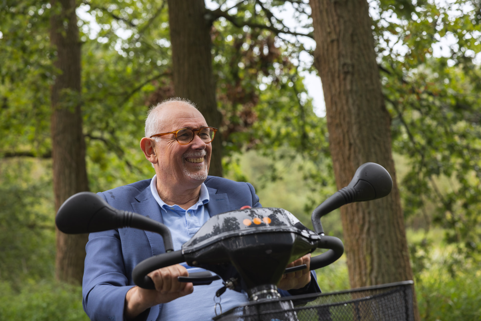 Een man rijdt glimlachend op een scootmobiel door een groen bos.